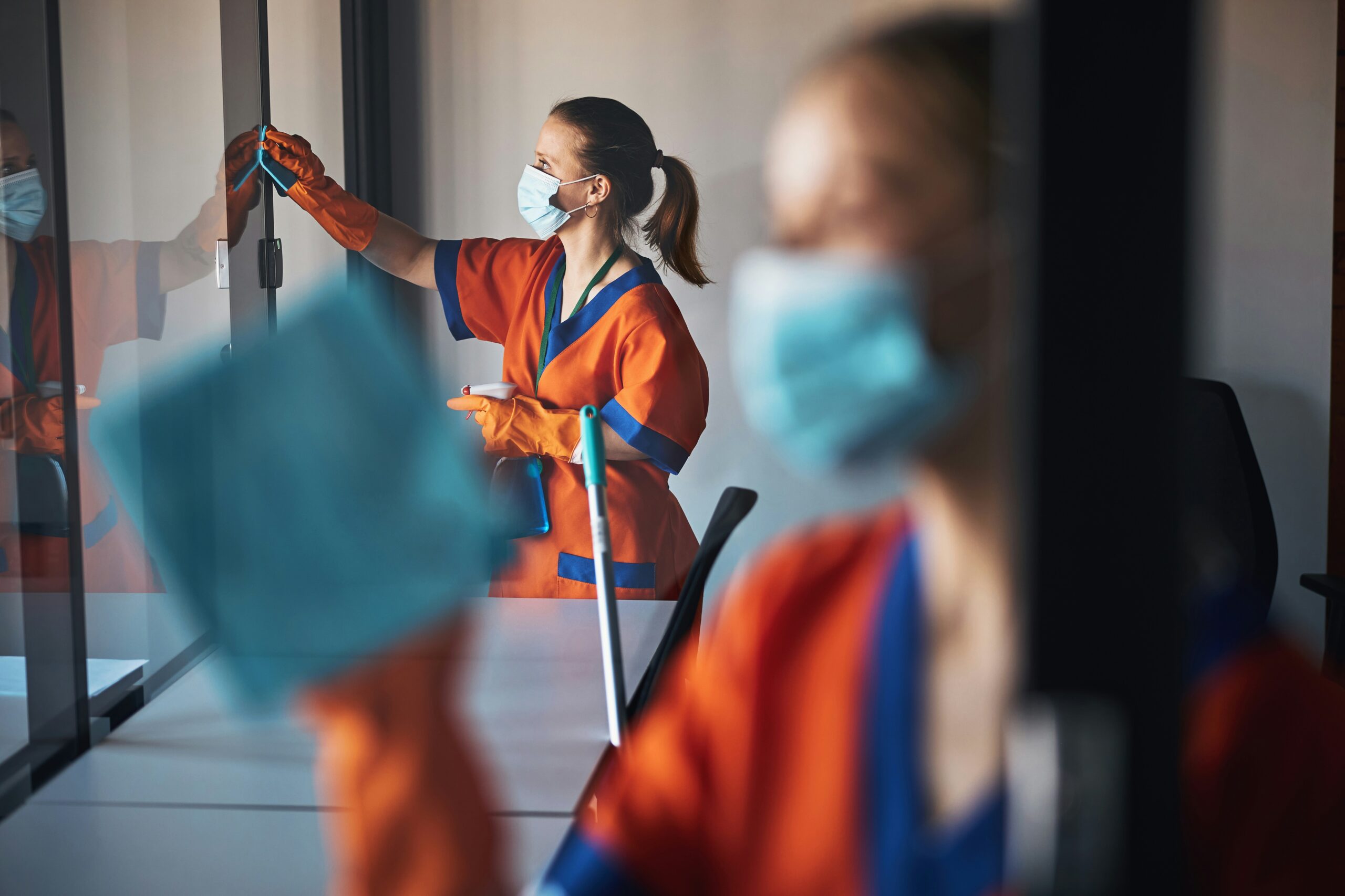 Zwei weibliche Reinigungskräfte tragen Einwegmasken und Handschuhe beim Reinigen von Glasscheiben.